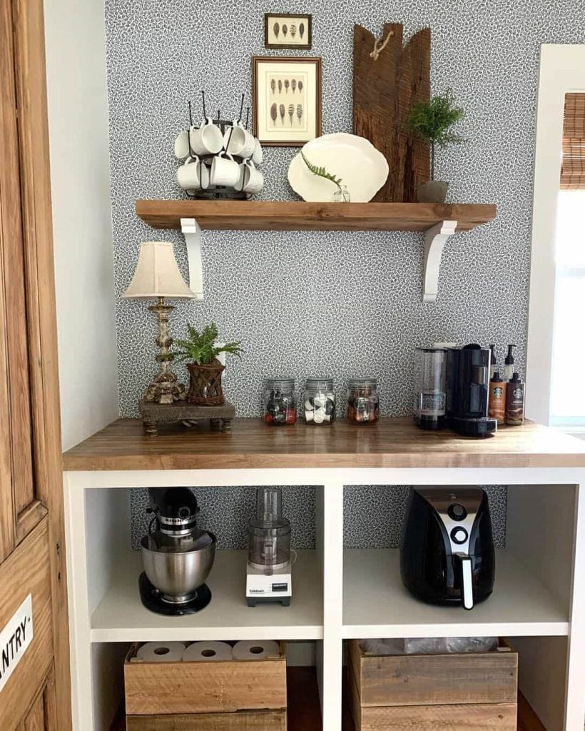 A home coffee bar featuring blue and white patterned wallpaper, wooden shelves, and white cabinetry. The setup includes a coffee machine, decorative items, and a stylish mug rack, creating a cozy and functional space.