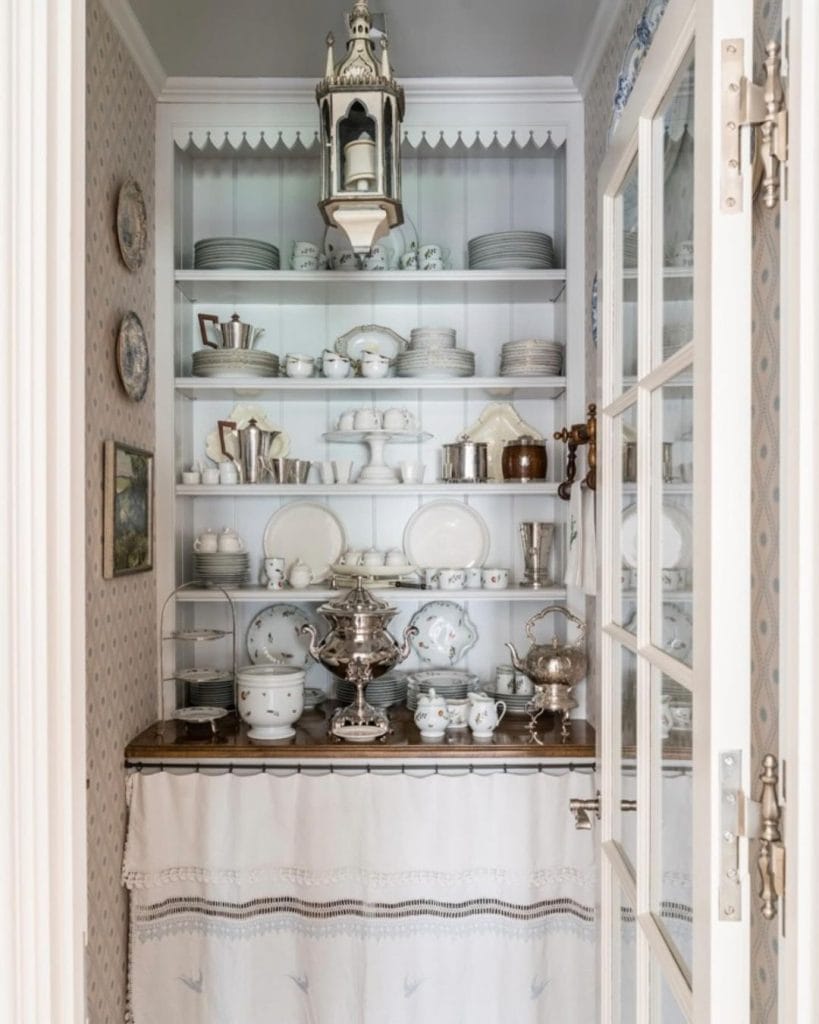 Vintage-style coffee nook with white shelving, fine china, antique silver pieces, and traditional lighting.