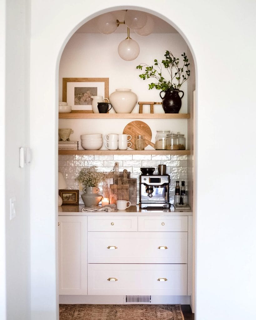 Stylish coffee bar set within an arched alcove, featuring white cabinetry, open wooden shelves, and soft lighting.