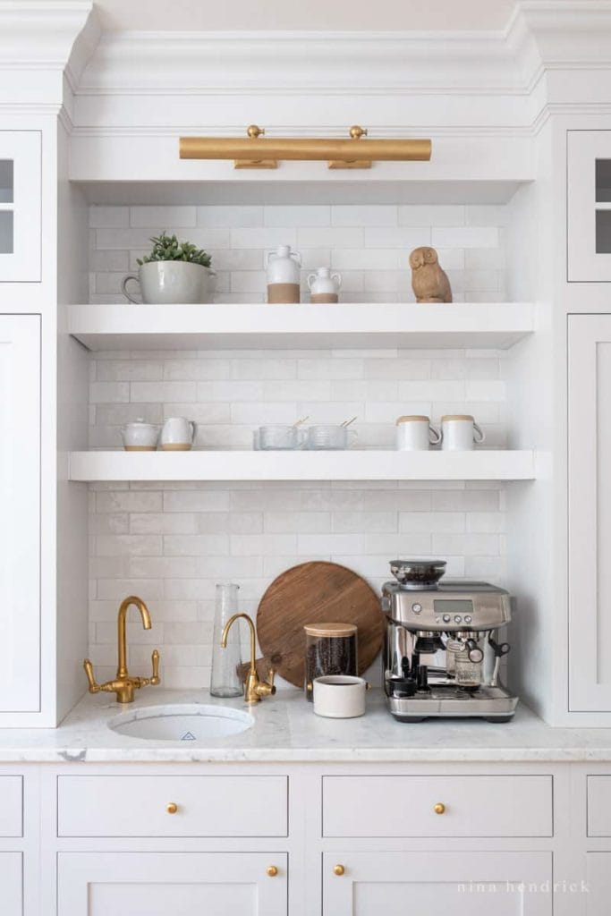 Elegant coffee bar with white cabinetry, gold fixtures, marble countertop, and curated decor items.