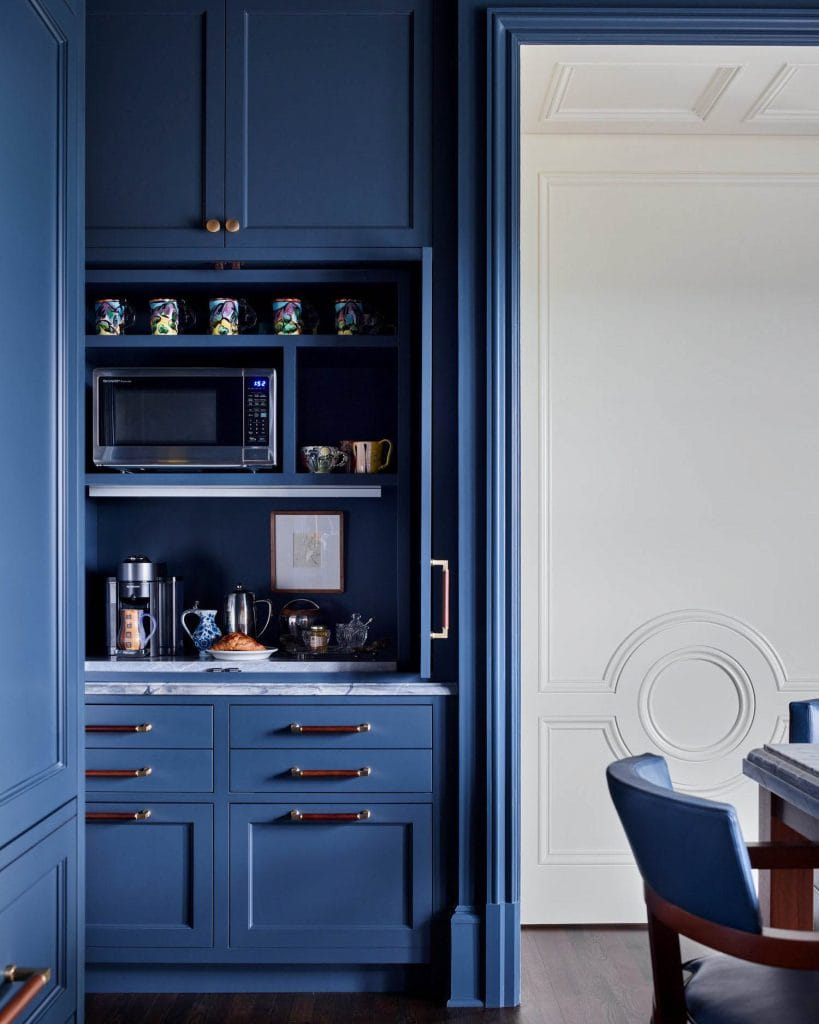 Bold blue coffee bar with navy cabinetry, marble countertop, gold hardware, and colorful mugs.