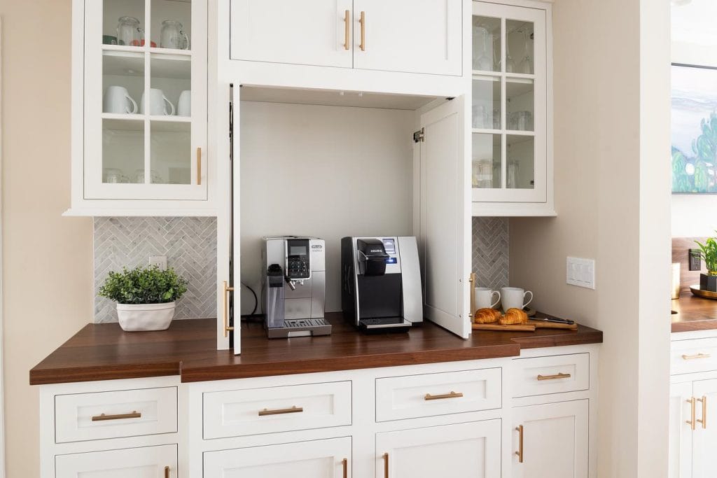 Countertop coffee station behind closed doors, white cabinetry, brass accents, and wooden countertop.