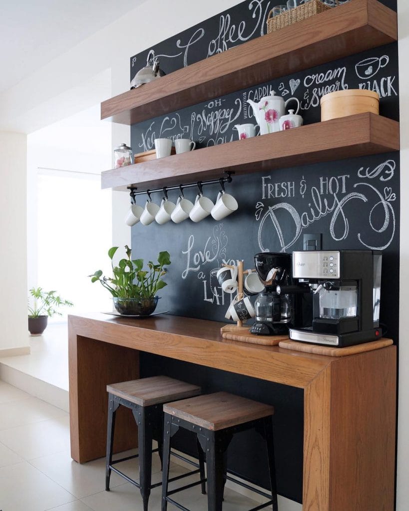 Coffee station with chalkboard wall, coffee-themed quotes, wooden counter, and shelves for coffee essentials.