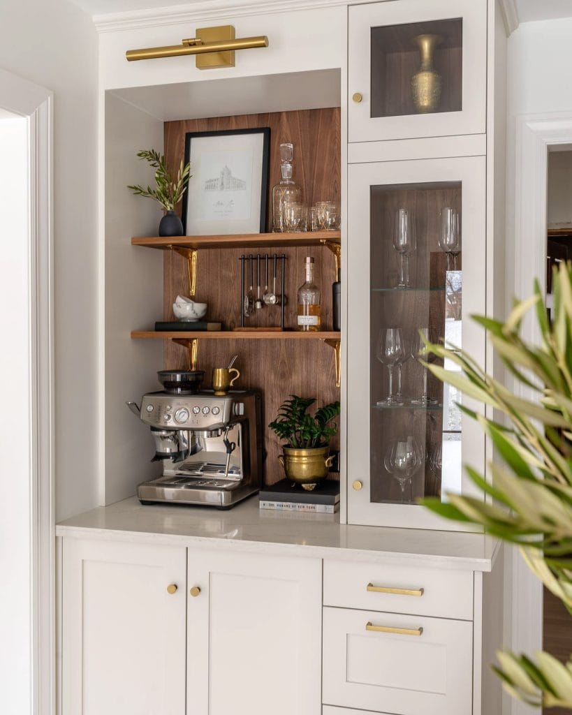 A coffee station with white cabinetry, walnut shelves, and brass accents. The espresso machine, greenery, and gold-toned decor create a luxurious and warm space.