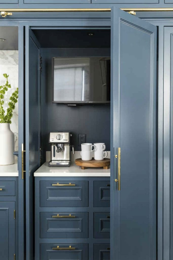 A hidden coffee station with dark blue cabinetry and brass hardware. An espresso machine and white mugs are neatly organized inside, providing a sleek and modern look.