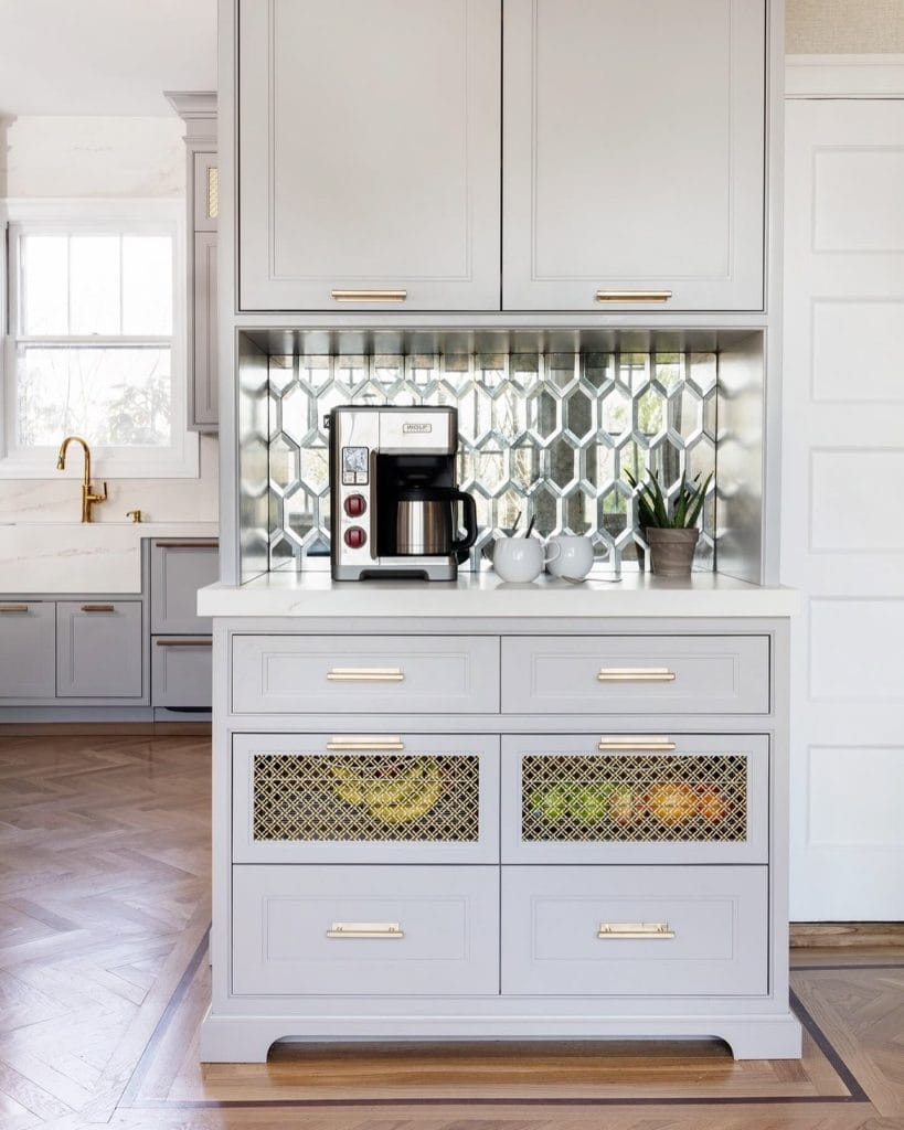 A coffee bar with light grey cabinets and a geometric glass tile backsplash. The space features a coffee maker and decorative items, creating a bright and stylish corner.