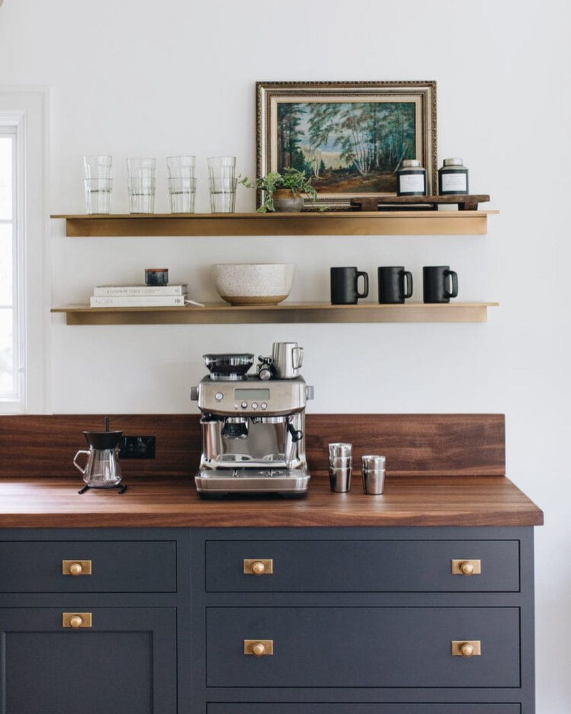 A coffee station with dark cabinetry, a wood countertop, and brass accents. Floating shelves display glassware and decorative items, adding to the sophisticated ambiance.