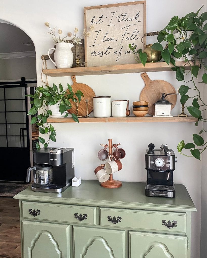 A rustic coffee station with a green vintage cabinet, wooden shelves, and plants. The setup includes an espresso machine, a coffee maker, and charming decor items for a cozy vibe.