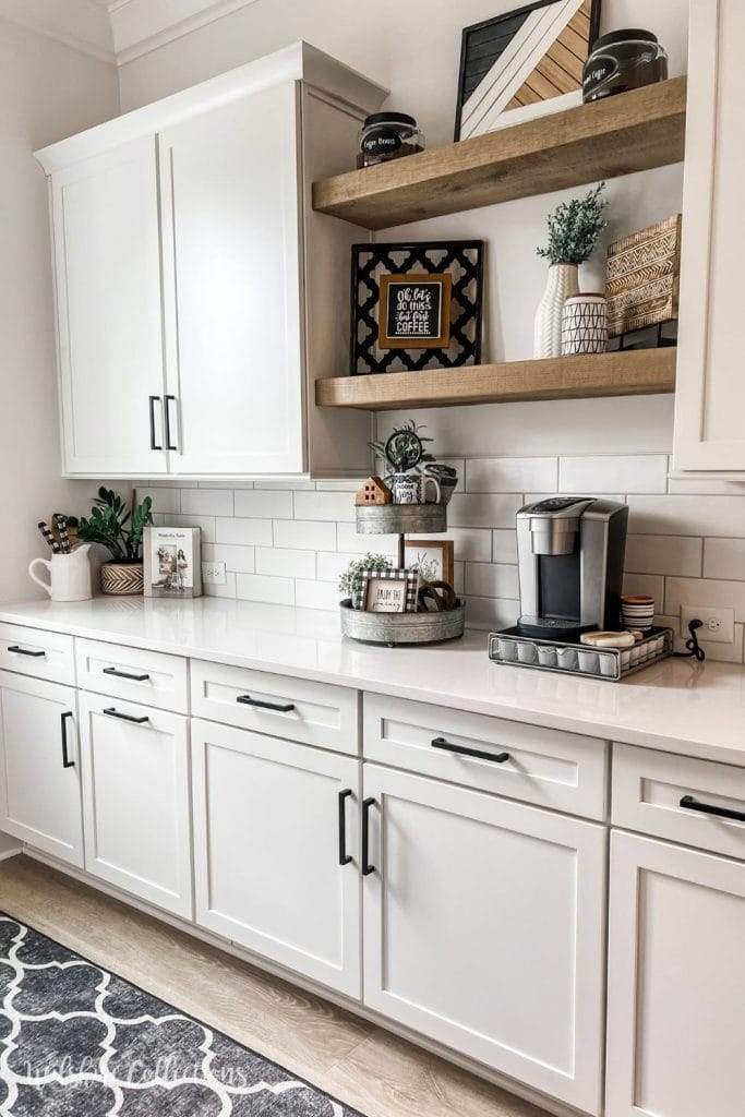 A modern farmhouse beverage station with white cabinetry and black handles, featuring wooden floating shelves adorned with decorative items and storage jars, and a sleek single-serve coffee machine on the countertop.