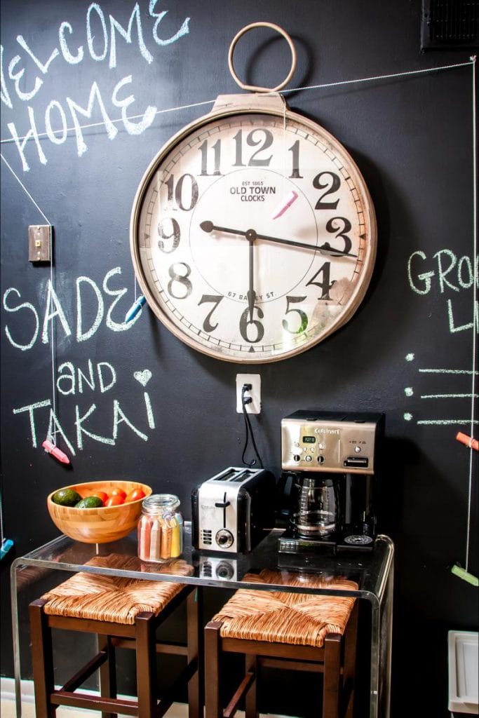 A coffee station with a large clock and a chalkboard wall with playful messages. The setup includes a coffee maker, toaster, and bar stools, creating a fun and vibrant corner.