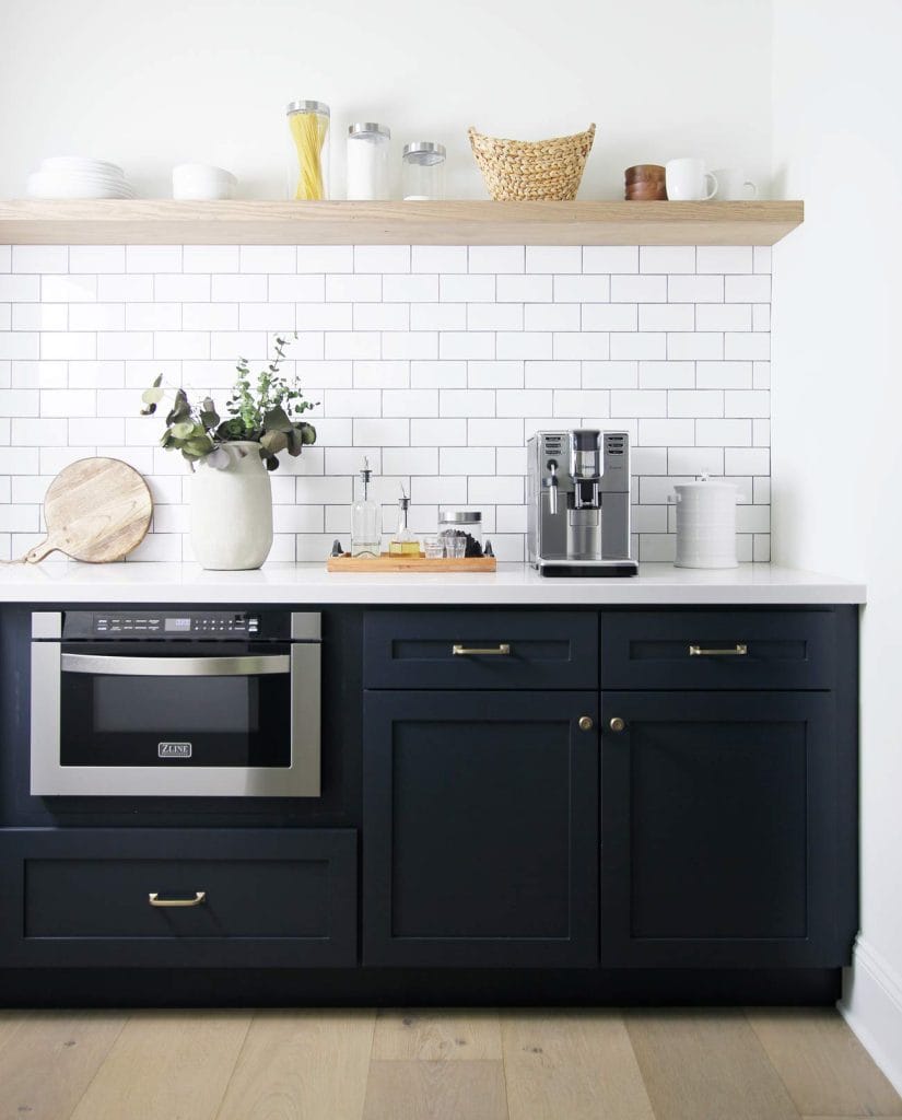 Elegant minimalist beverage station with dark navy cabinetry, white countertop, and classic subway tile backsplash, featuring open wooden shelves with kitchen essentials and decorative items.