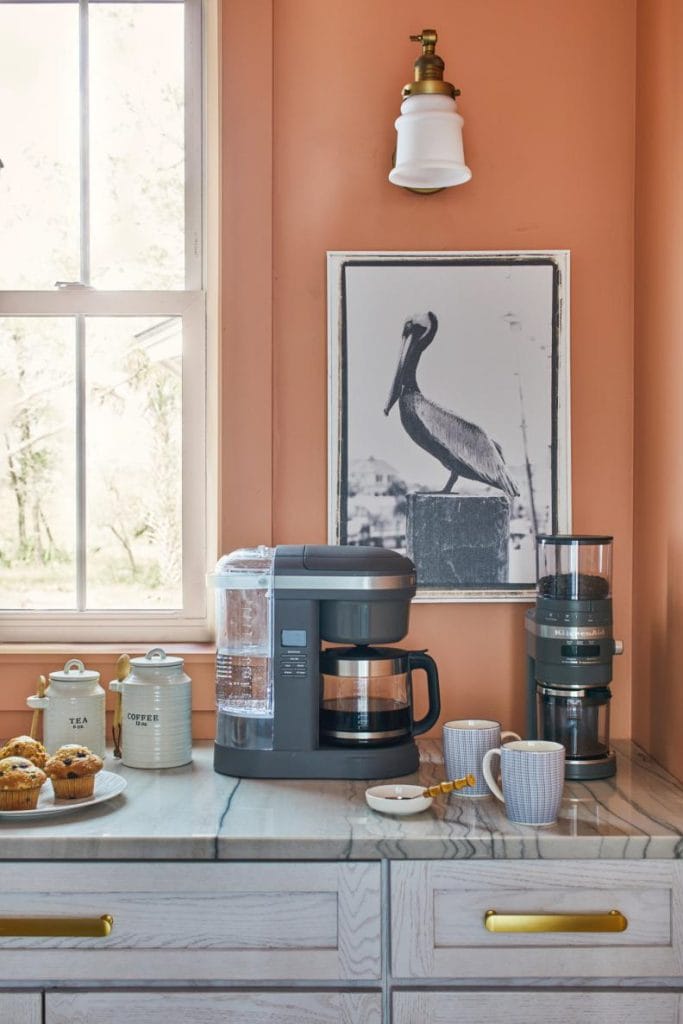 A coral-colored wall with matching artwork and a modern coffee maker, paired with light wood cabinetry and a marble countertop.