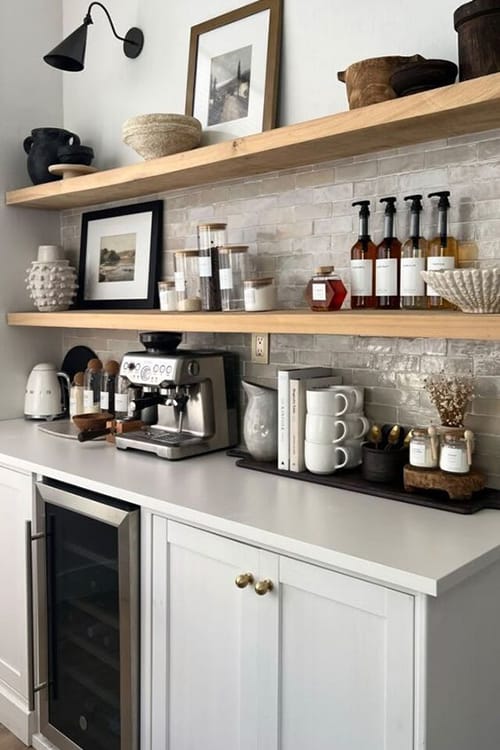 A modern home beverage station featuring white cabinetry with light wooden shelves, adorned with coffee syrups, mugs, and decorative items. The setup includes a sleek espresso machine, a built-in mini fridge, and minimalist decor against a textured tile backsplash.