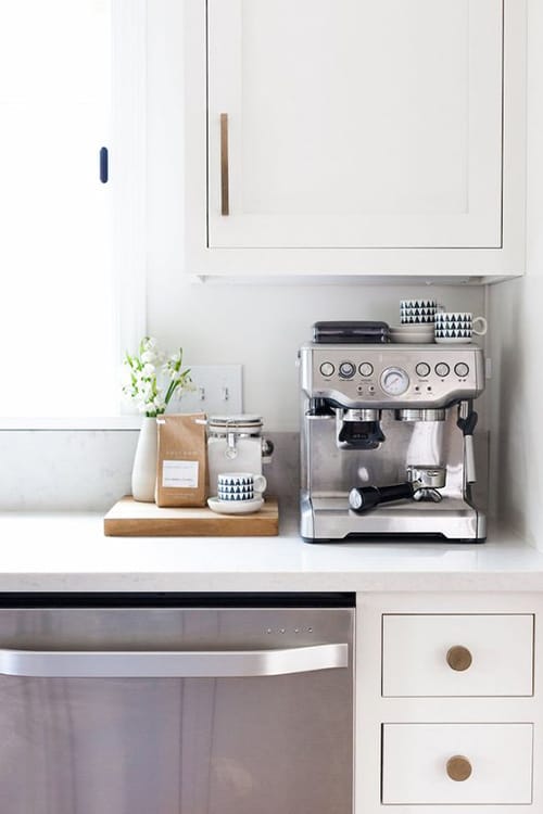 A sleek coffee bar with white cabinetry, a stainless-steel coffee maker, and minimalist decor.