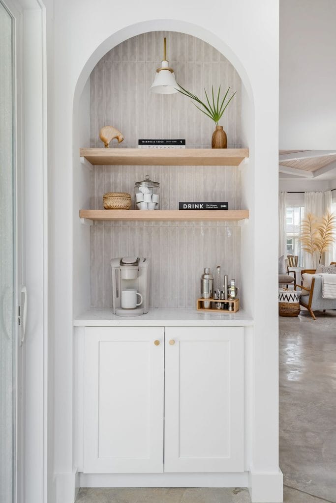 An arched nook home bar with white cabinetry, a tiled backsplash, and wooden shelves. The shelves hold a small potted plant, wicker basket, and bar tools, creating a sleek and minimalist design.