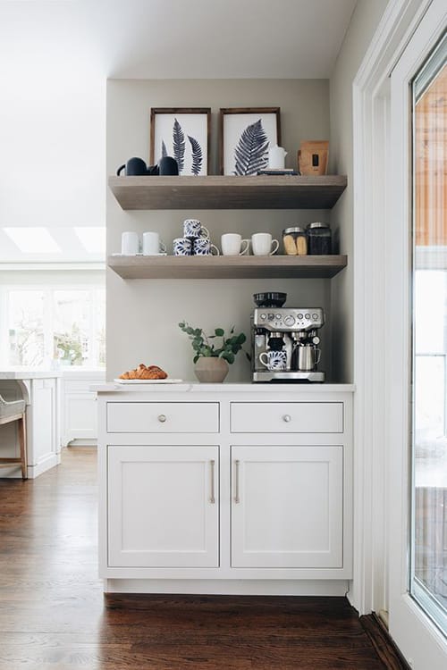 A sleek, modern kitchen setup featuring dark cabinets and light wood floating shelves with an espresso machine, mugs, and other kitchenware against a light gray tiled backsplash.