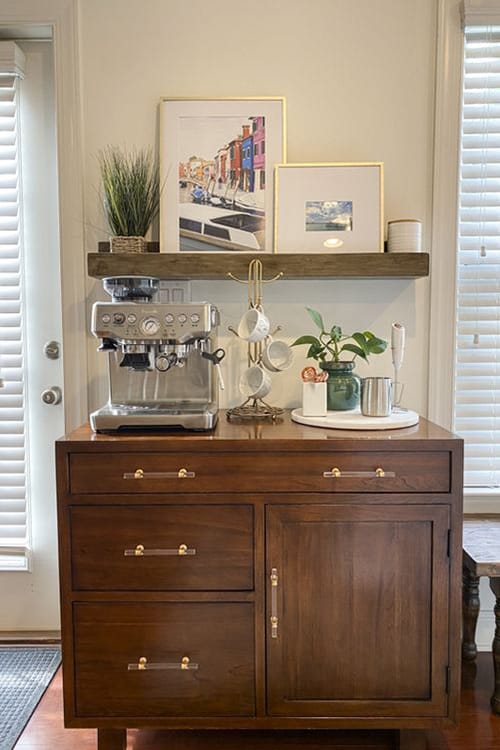 A sleek, modern kitchen setup featuring dark cabinets and light wood floating shelves with an espresso machine, mugs, and other kitchenware against a light gray tiled backsplash.