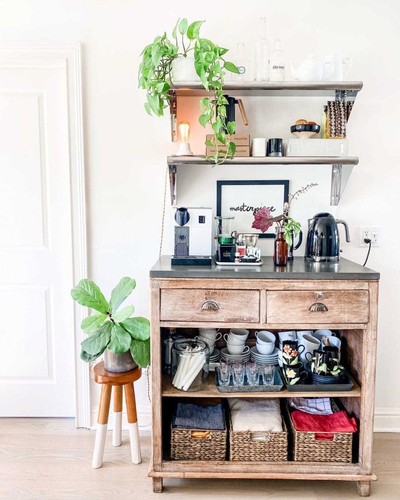 A rustic coffee bar with a weathered wooden cabinet, open shelving, and greenery, featuring baskets and jars for storage and decor.