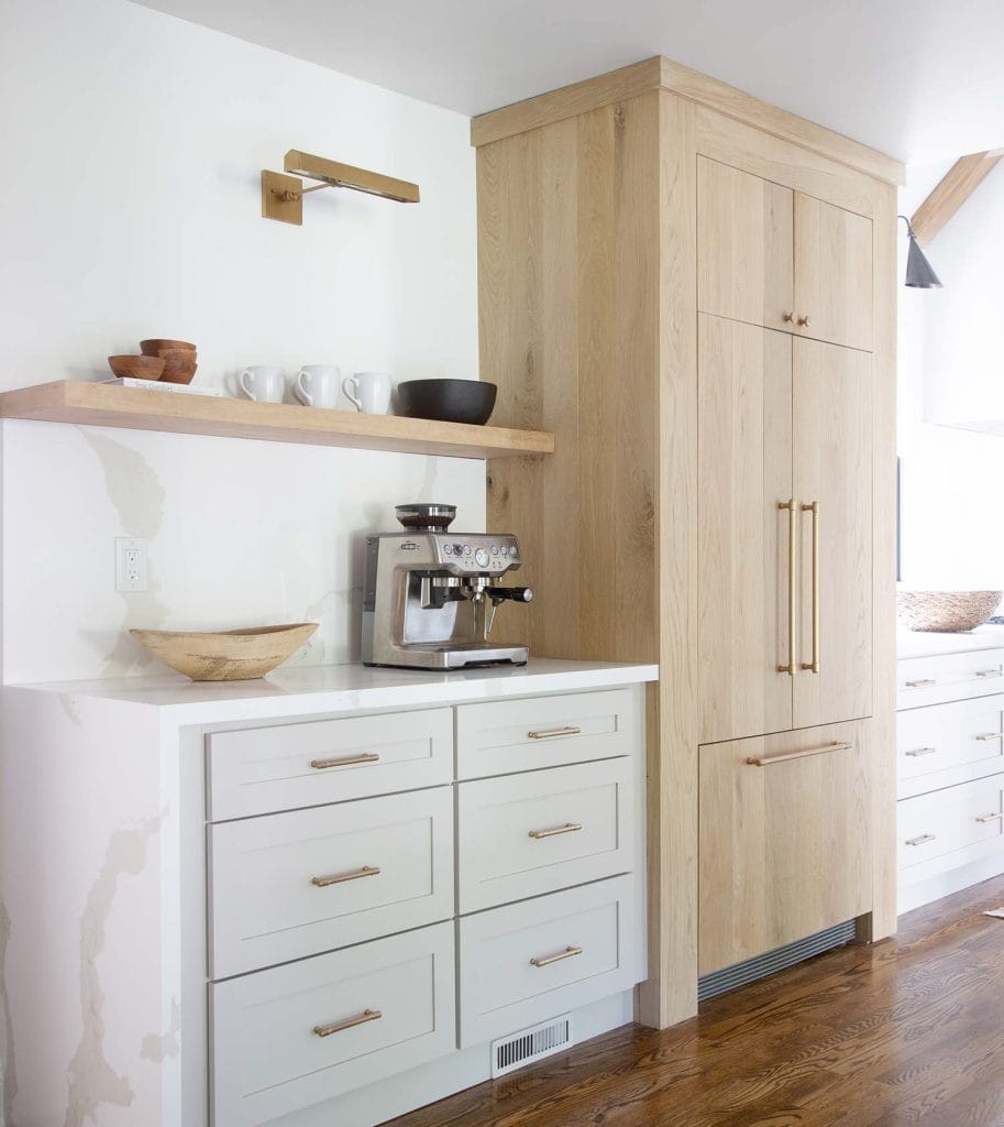 This minimalist coffee bar features sleek white cabinetry with brass hardware, complemented by a light wood cabinet and floating shelf. The design is clean and modern, creating a stylish and functional space for coffee preparation.