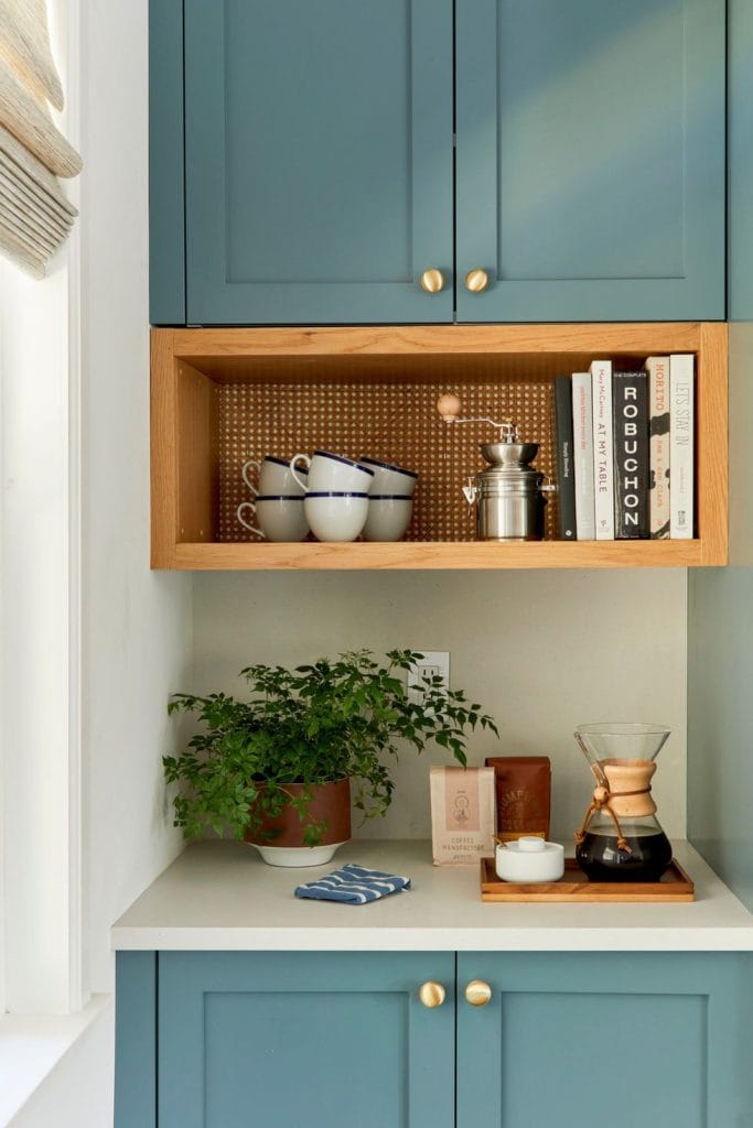 A cozy home coffee bar featuring teal cabinetry with gold hardware, a wooden open shelf with a textured backdrop, a potted plant, a Chemex coffee maker, and neatly stacked coffee bags on the countertop.