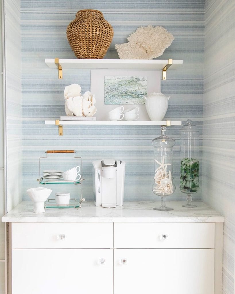 Coastal-themed coffee bar with white cabinetry, marble countertop, soft blue wallpaper, and beach-inspired decor elements.