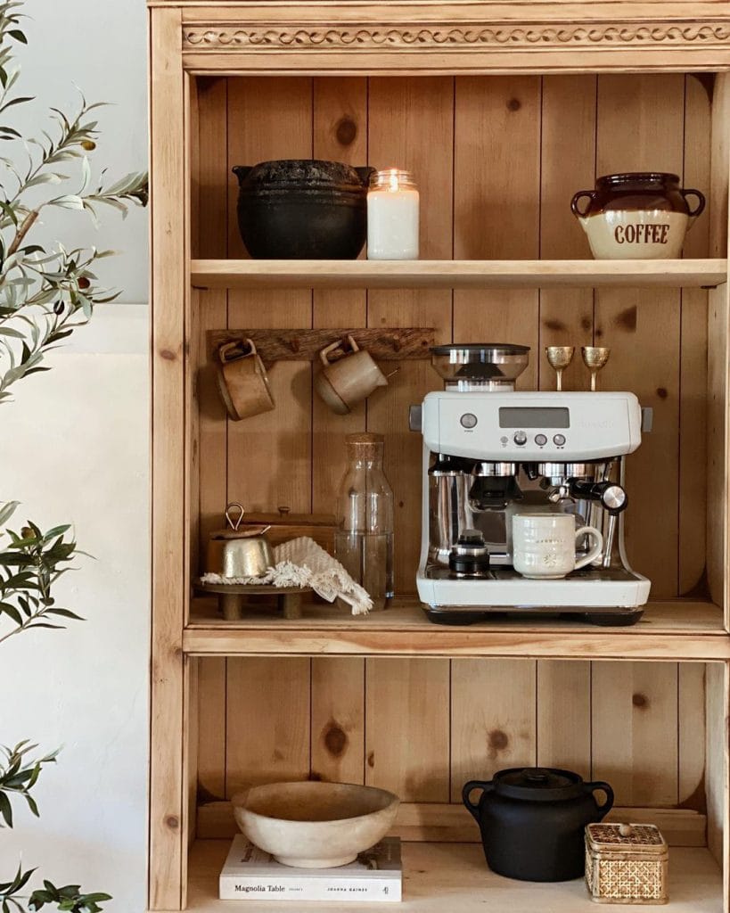 Rustic wooden shelf unit with intricate carved details, displaying a coffee machine, ceramic pot, candle, and various vintage mugs, creating a cozy and nostalgic ambiance.