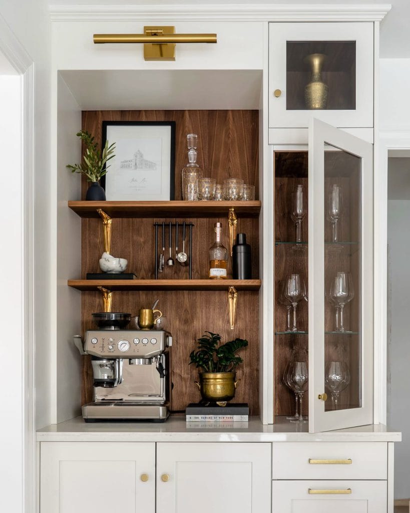 A sophisticated home beverage bar with white cabinetry, a warm wooden backdrop, and brass accents. The setup includes a professional espresso machine, glassware, and decorative items, creating a chic and functional space.