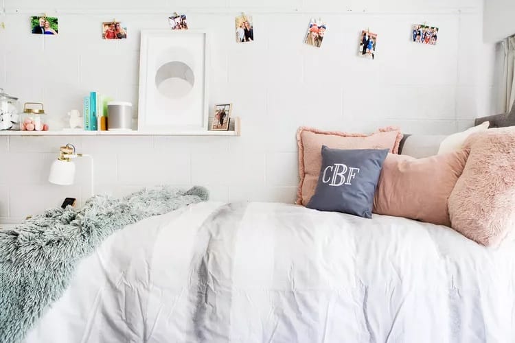 A cozy dorm room bed with plush pillows, monogrammed cushion, and a shelf above displaying photos and decor items against white walls.