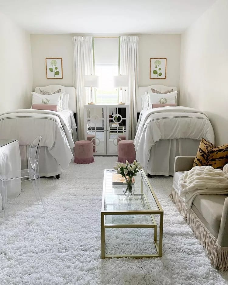 A dorm room with twin beds, white bedding, gold accents, clear chairs, a fluffy white rug, and soft pink decor elements.