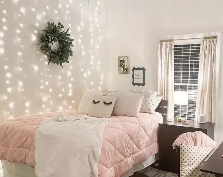 A dorm room with a bed covered in pink bedding, a wall decorated with string lights, and cozy decor elements.