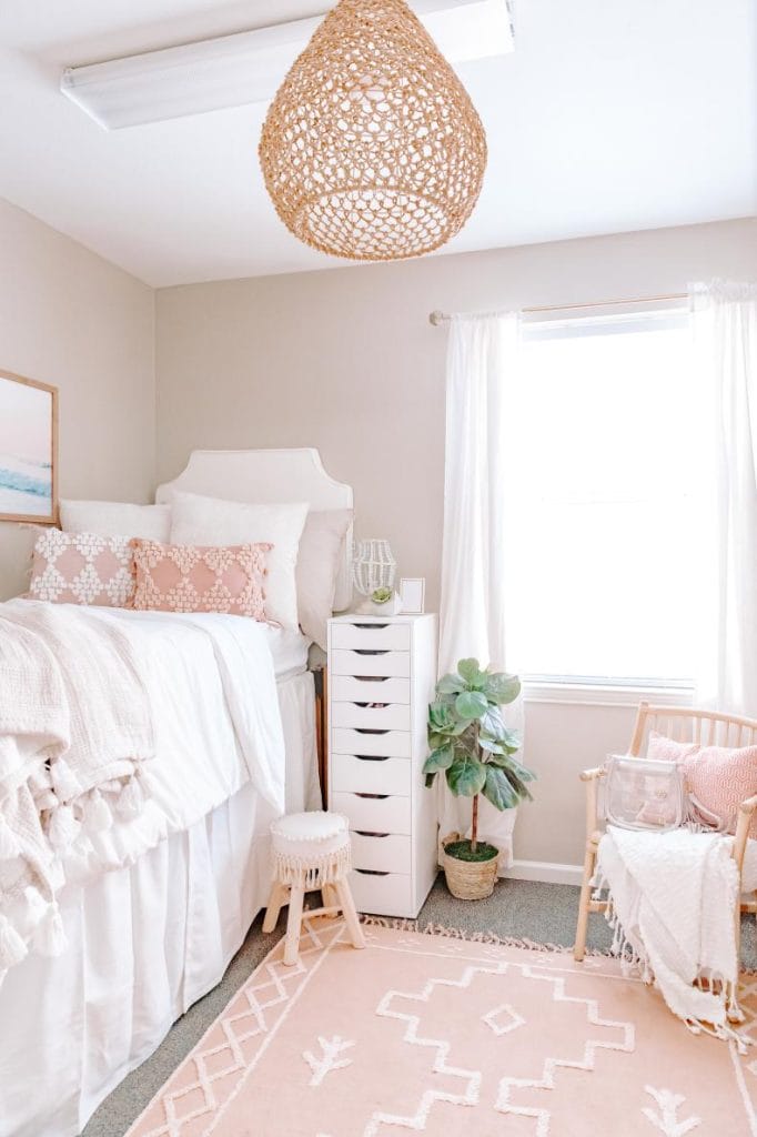  Cozy pink and white dorm room with soft pink accents, woven light fixture, and potted plants.