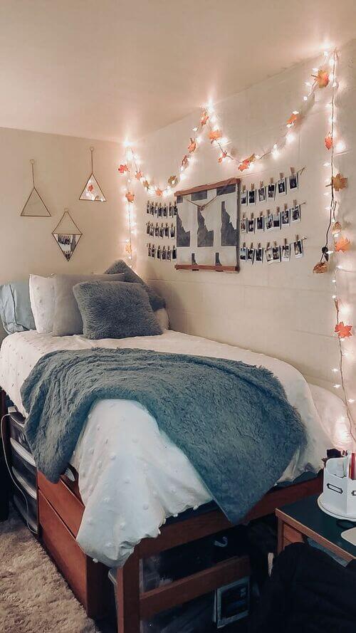 A dorm room with fall-themed string lights, leaf garlands, plush bedding, and a personal photo wall.