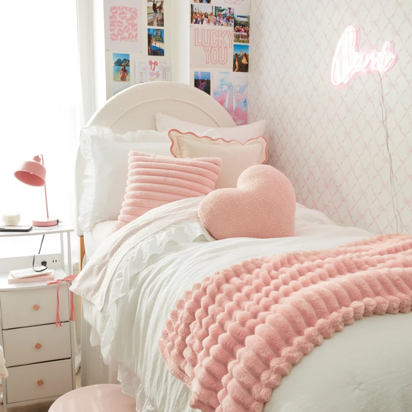 Feminine dorm room with pink and white bedding, textured pillows, neon sign, and a photo collage creating a trendy vibe.