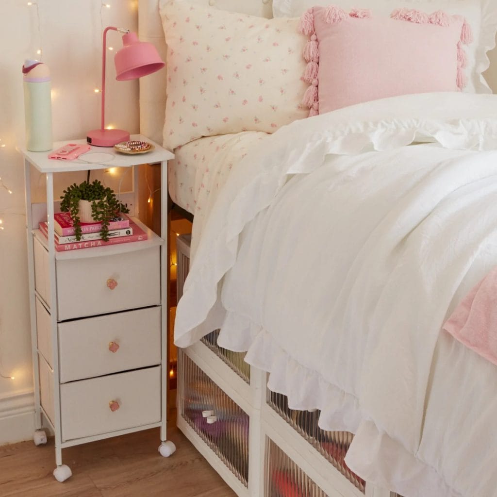 A bed with pink and floral accents, including a pink lamp and decorative books on a nightstand, with soft, ruffled bedding.