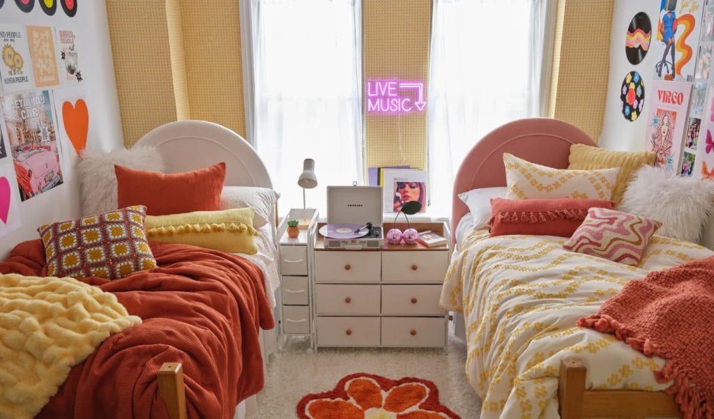 A vibrant room with two beds, retro bedding, colorful pillows, a shared nightstand with a record player, and neon wall decor.