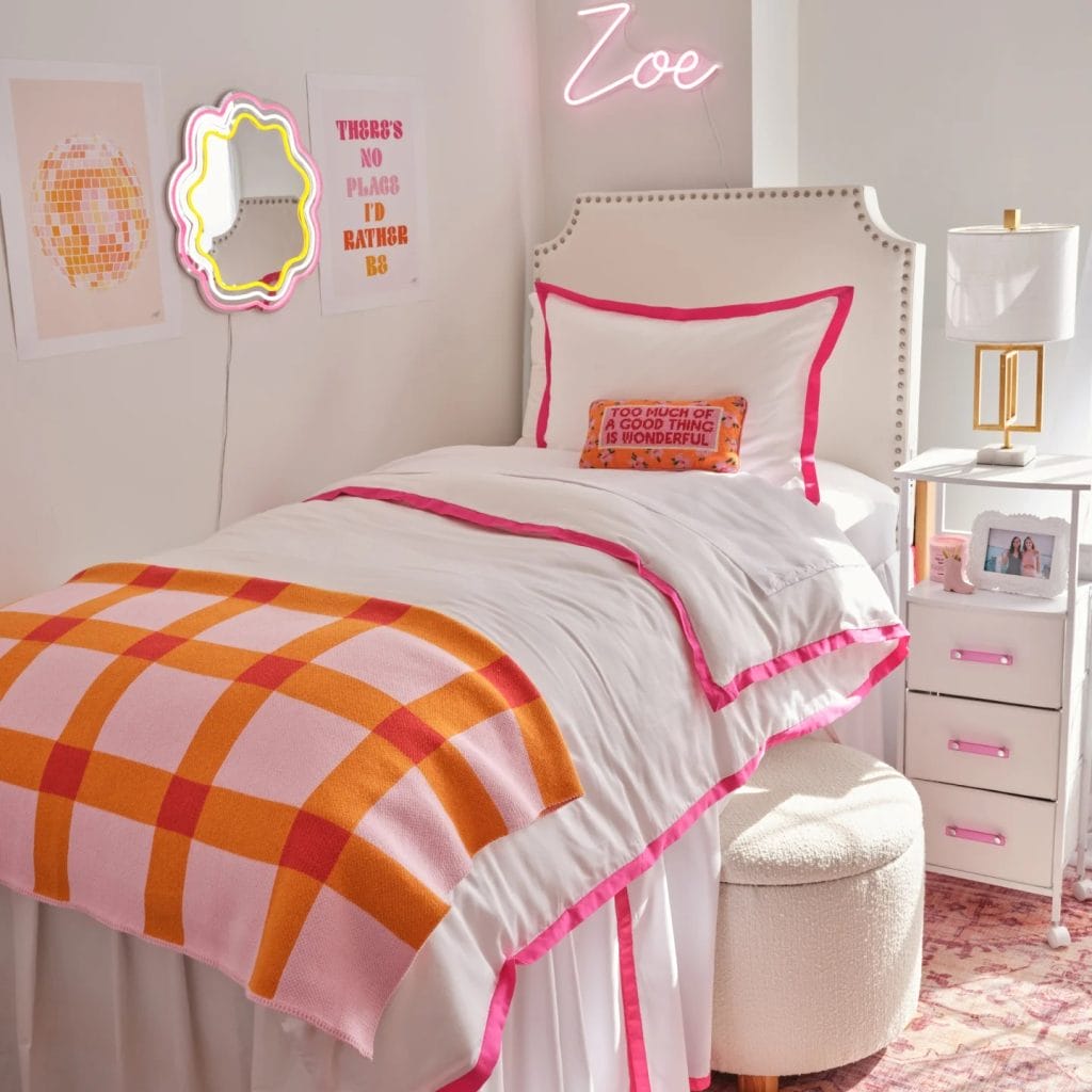 A room with a white bed, pink and orange bedding, neon name sign, colorful wall art, and a stylish nightstand.