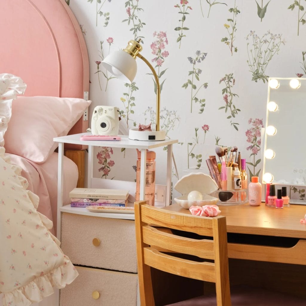 A feminine workspace with a wooden desk, makeup vanity, floral wallpaper, soft pink bedding, and a gold lamp.