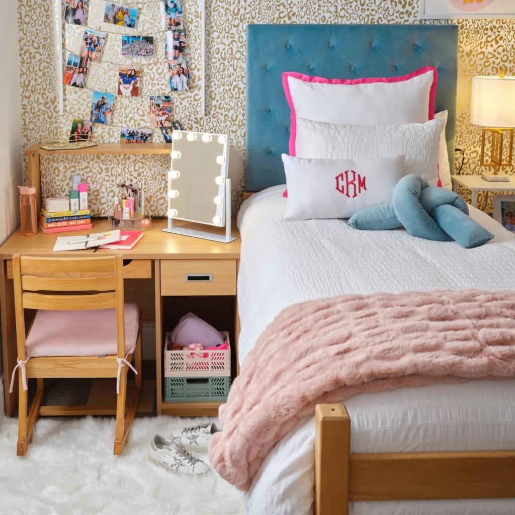 A stylish room with a blue tufted headboard, white and pink bedding, a cozy desk area with a lighted vanity mirror, and a photo wall display.