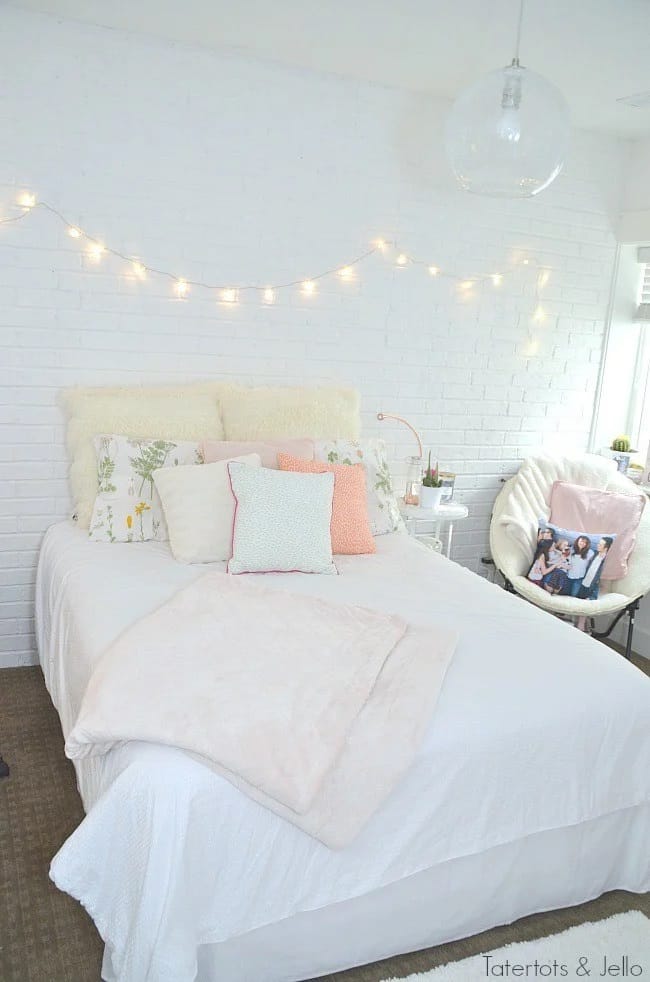 A bedroom with white bedding, pastel pillows, and string lights creating a cozy atmosphere.