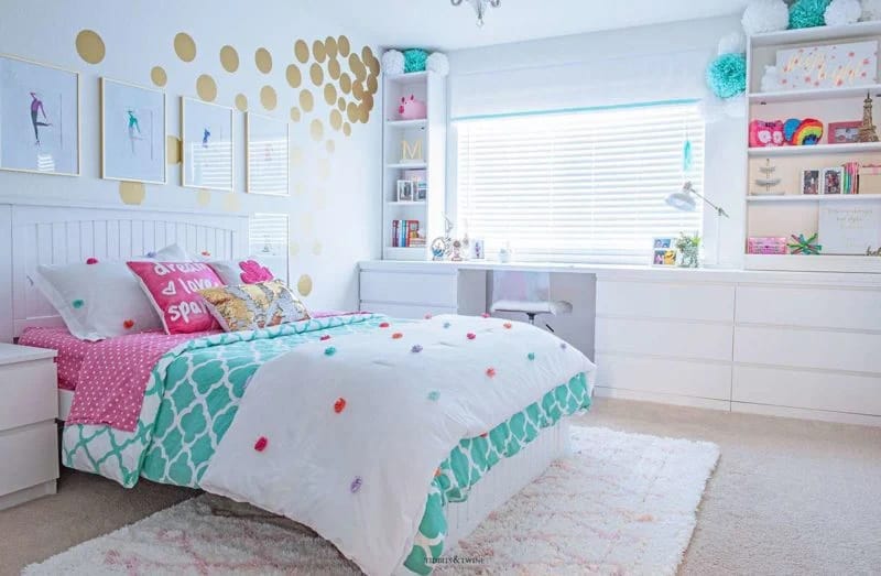 A white bed with colorful pom-poms and vibrant pillows in a room with a gold polka-dot wall and a study desk.