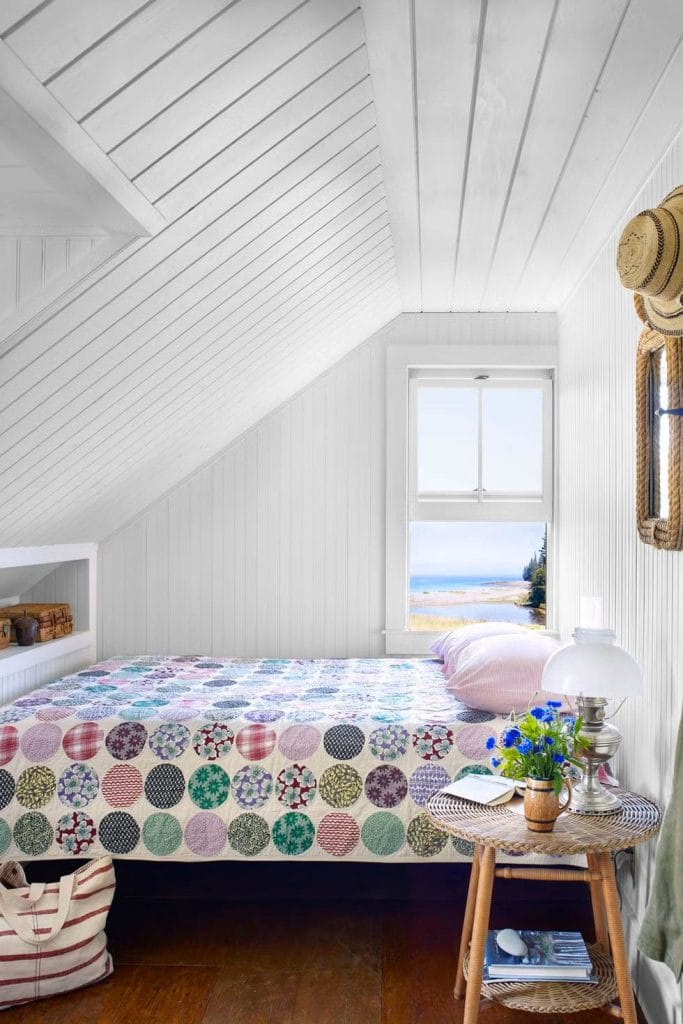 Attic bedroom with white paneled walls, a colorful patchwork quilt, and a coastal view.