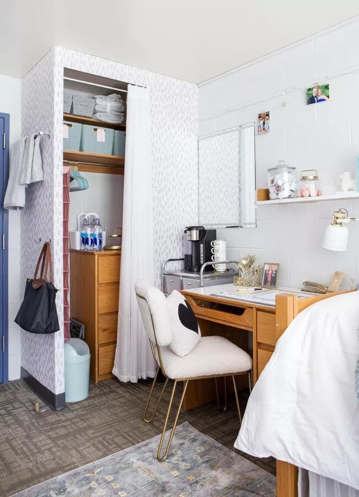 An organized dorm room corner with a wooden desk, stylish chair, curtained closet, and various storage solutions including bins and shelves.