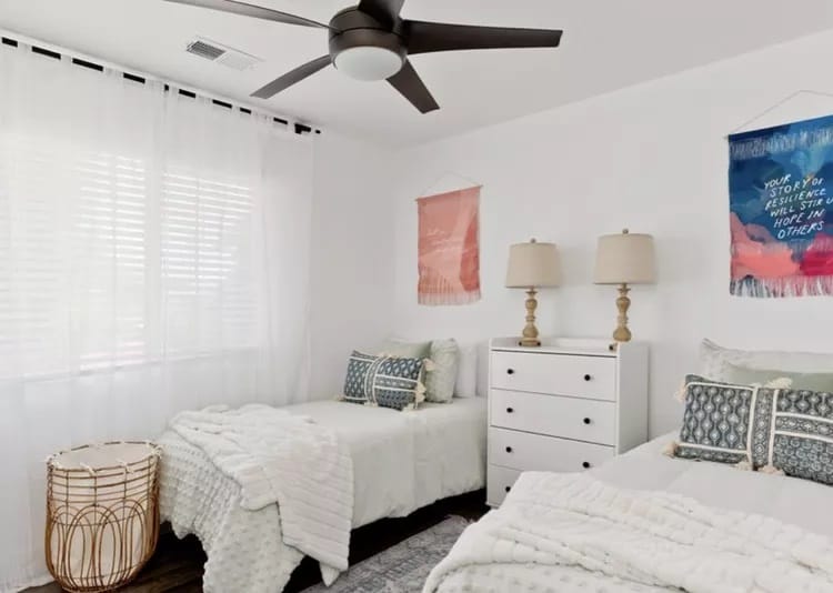 A dorm room with twin beds, white bedding, blue accent pillows, large windows with sheer curtains, a ceiling fan, and simple wall art.