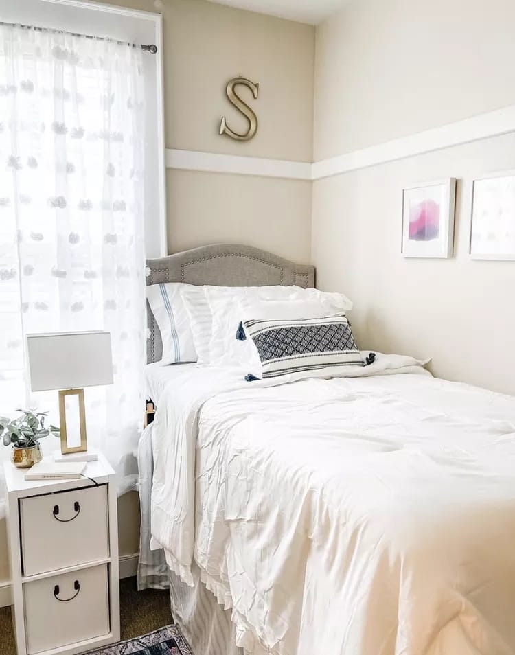 A dorm room with a single bed, white bedding, a small bedside table with a lamp, and minimal wall decor.