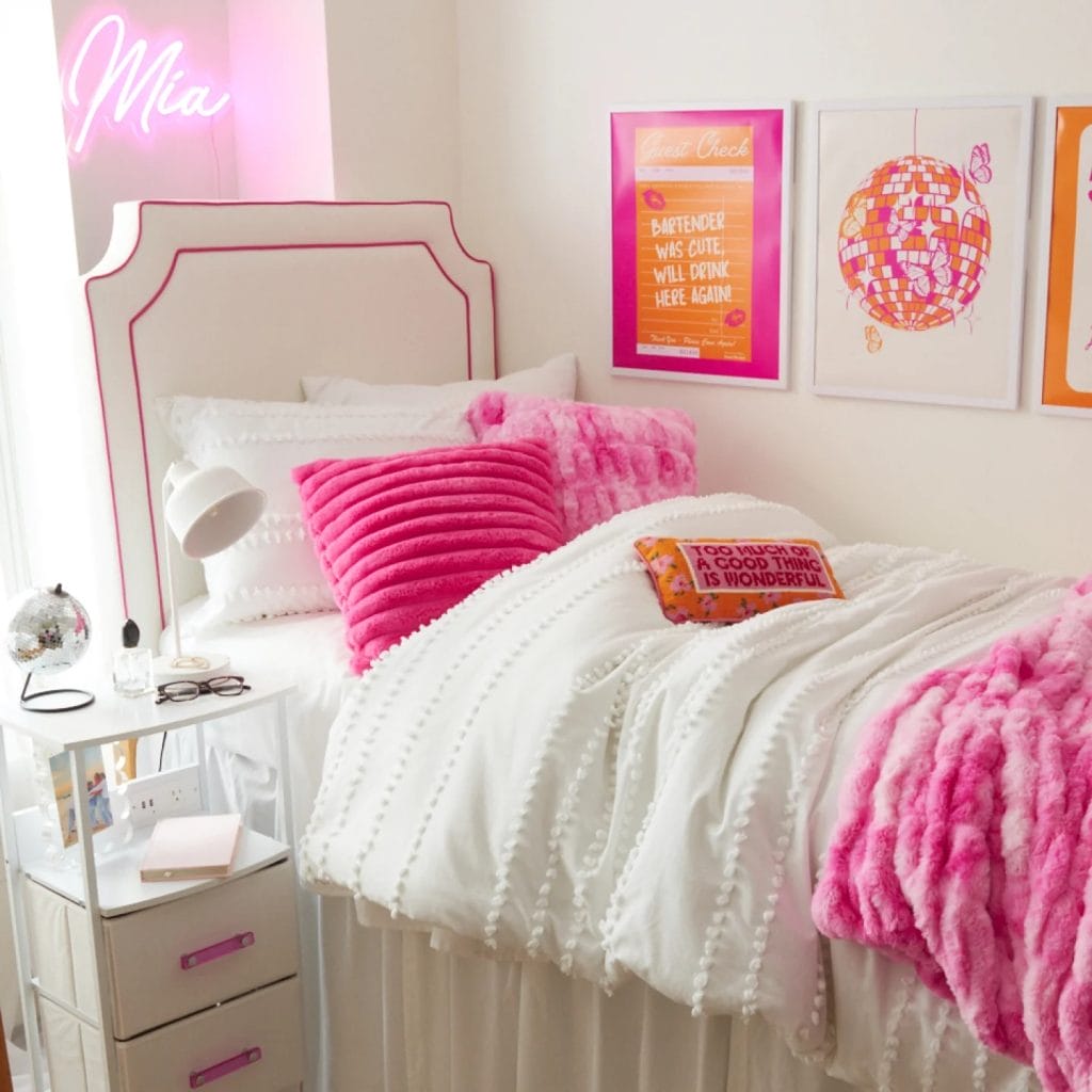 A bed with vibrant pink pillows and blanket, neon name sign, and quirky wall art.