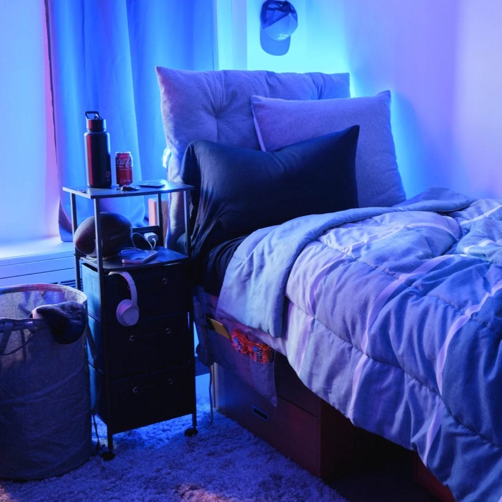 A cozy room with blue lighting, featuring a bed with grey and black pillows, a bedside table holding a water bottle and soda can, and under-bed storage.