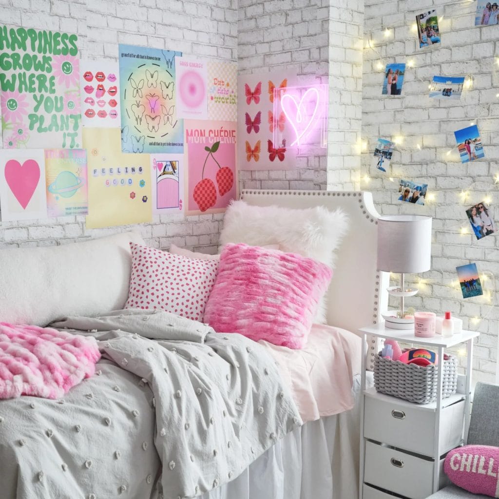 A cozy room with a white bed, pink pillows, positive-themed wall art, a neon heart sign, and string lights.