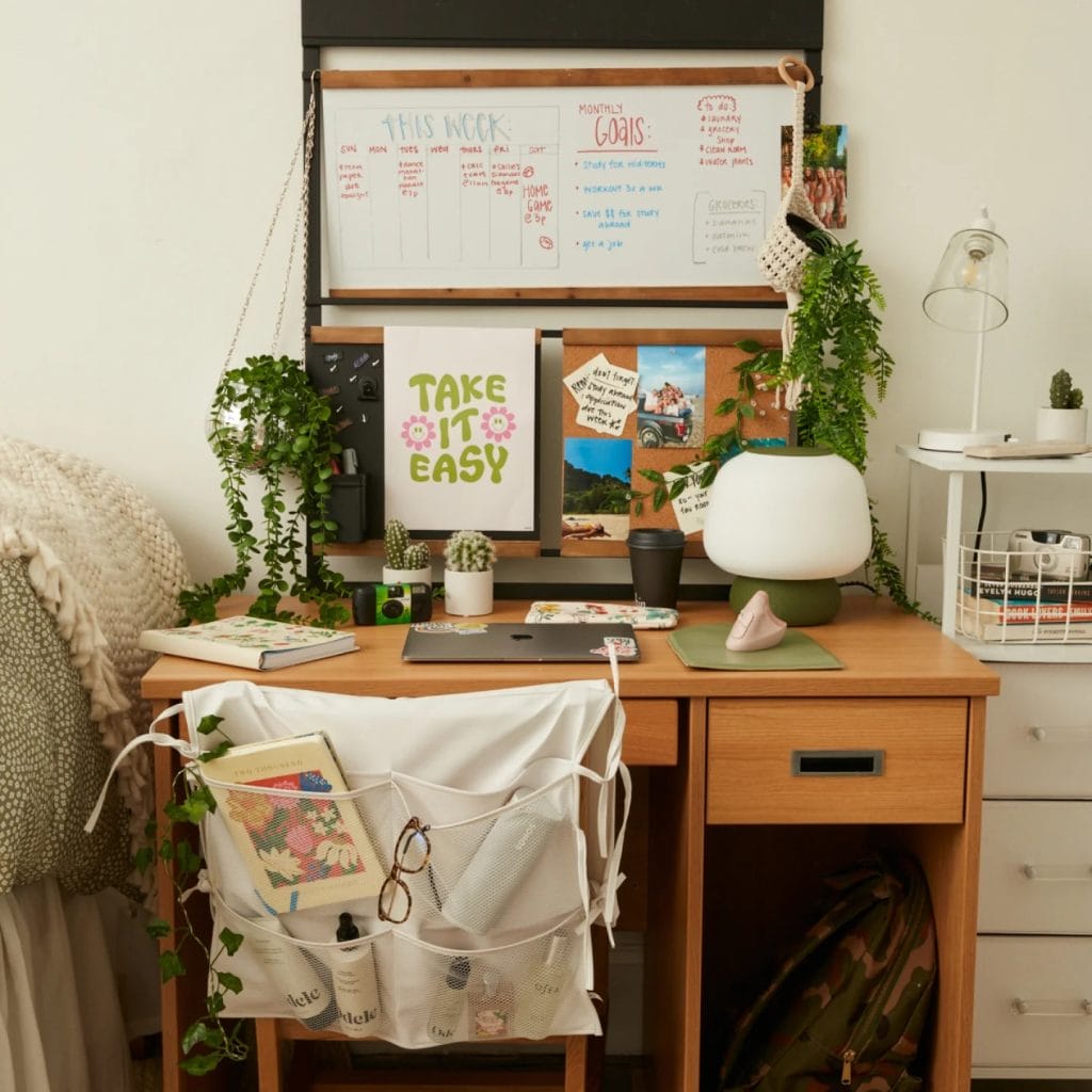 Dorm room study nook with wooden desk, hanging plants, whiteboard, and bulletin board.