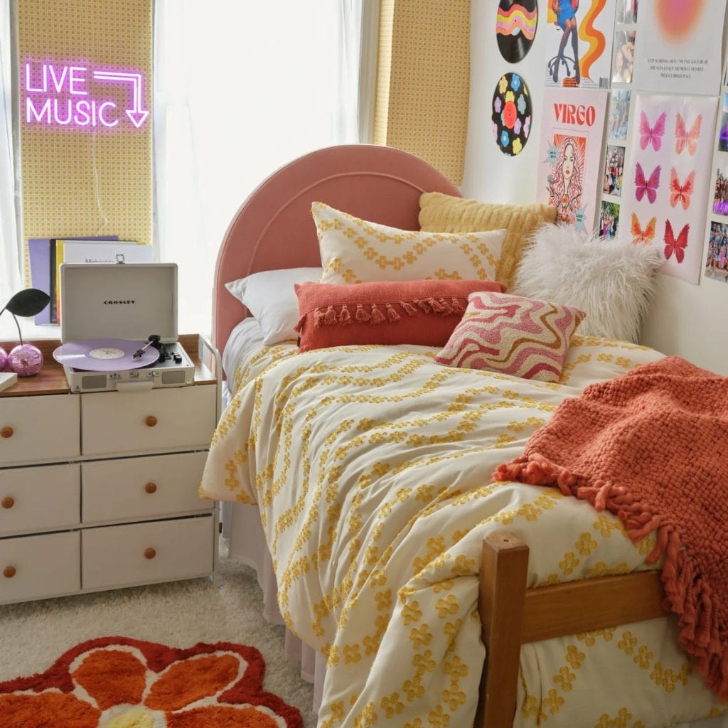 A sunny room with a yellow and white bedspread, orange and pink pillows, and playful wall decor including a neon sign and record art.