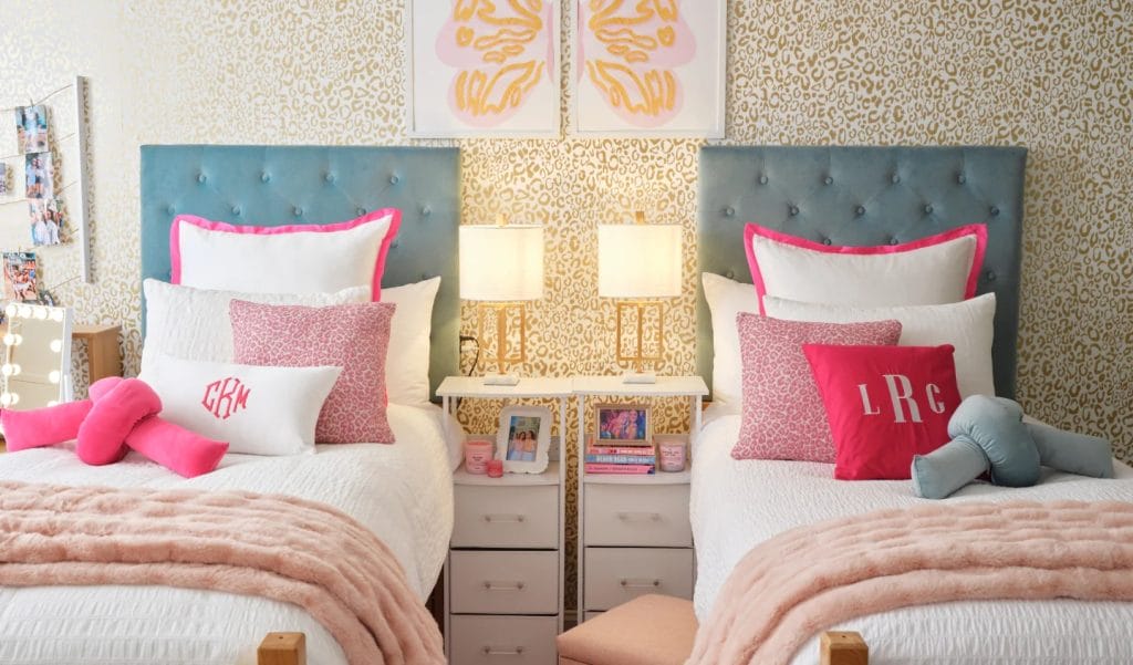 A chic room with twin beds, white bedding, pink accents, tufted headboards, gold decor, and a shared nightstand.